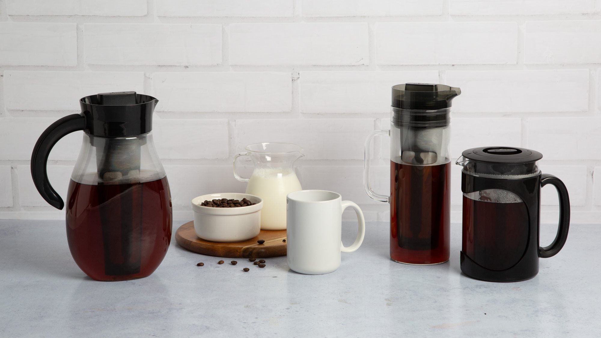 Primula cold brew carafes and coffee press with beans, milk and a mug against white brick